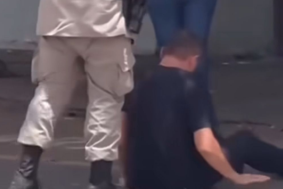 Imagem colorida mostra um homem de camisa escura sentado no chão ao lado de um policial militar.