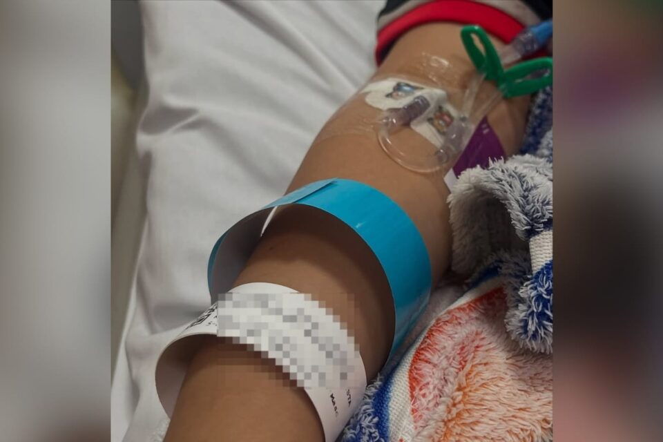 A imagem mostra uma foto do braço da criança com acessos para soro e pulseirinha hospitalar