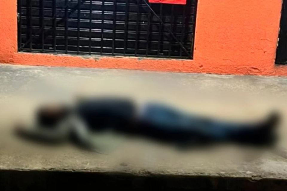Imagem colorida mostra um homem caído na calçada de uma rua em frente a distribuidora com a parede laranja e a porta de ferro abaixada.