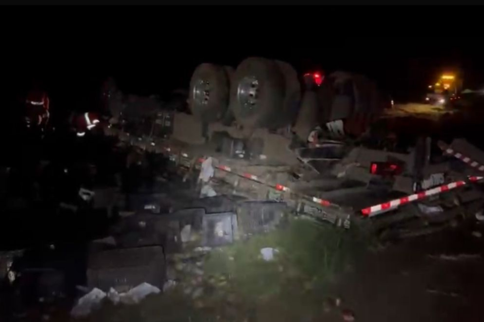 Imagem colorida mostra um caminhão tombado na beira de uma rodovia a noite.