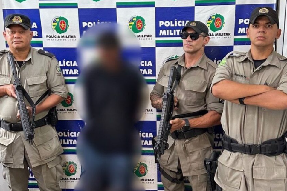 Imagem colorida mostra um homem com uma blusa e uma calça jeans azul no meio de três policiais após ser preso.