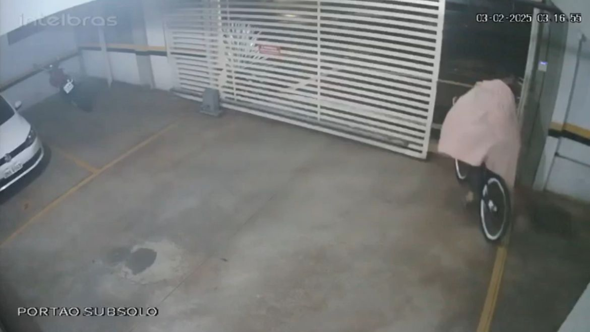 Imagem colorida mostra um homem usando uma coberta. Ele está em cima de uma bicicleta saindo de um estacionamento.