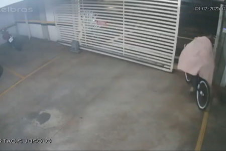 Imagem colorida mostra um homem usando uma coberta. Ele está em cima de uma bicicleta saindo de um estacionamento.