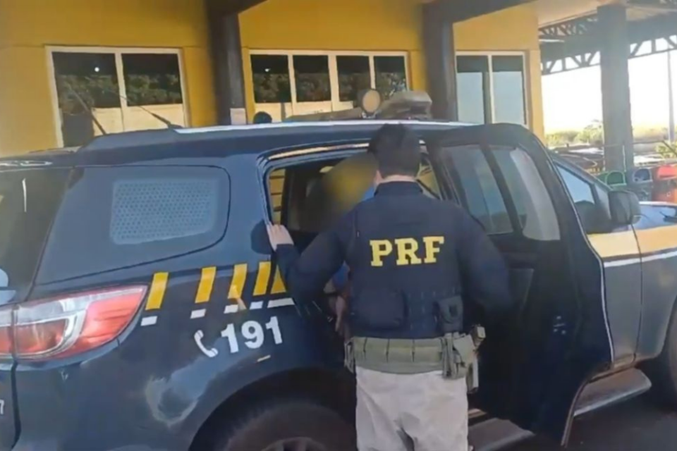 Imagem colorida mostra um agente d PRF ao lado da porta traseira da viatura.
