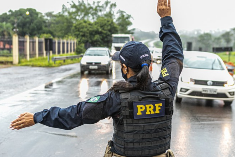 Imagem colorida mostra uma agente da PRF de costas com o braço direito levantado e o esquerdo apontando para o lado em uma rodovia.