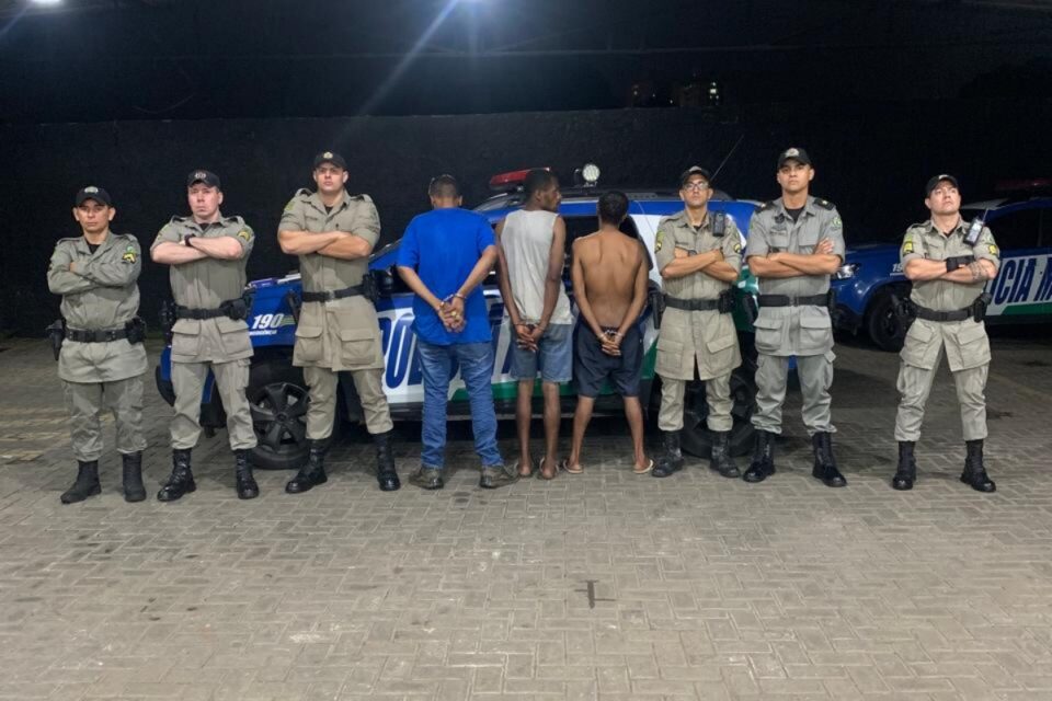 A imagem mostra os três suspeitos presos em frente a uma viatura da PM