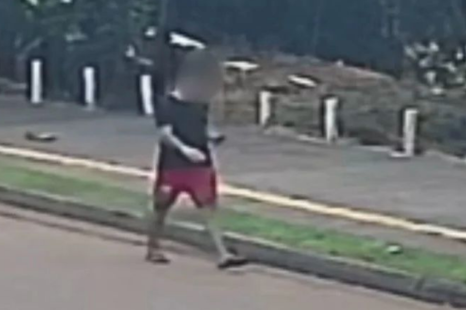 Imagem colorida mostra um homem andando pela rua. Ele usa uma camiseta preta, uma bermuda vermelha e um chinelo preto.