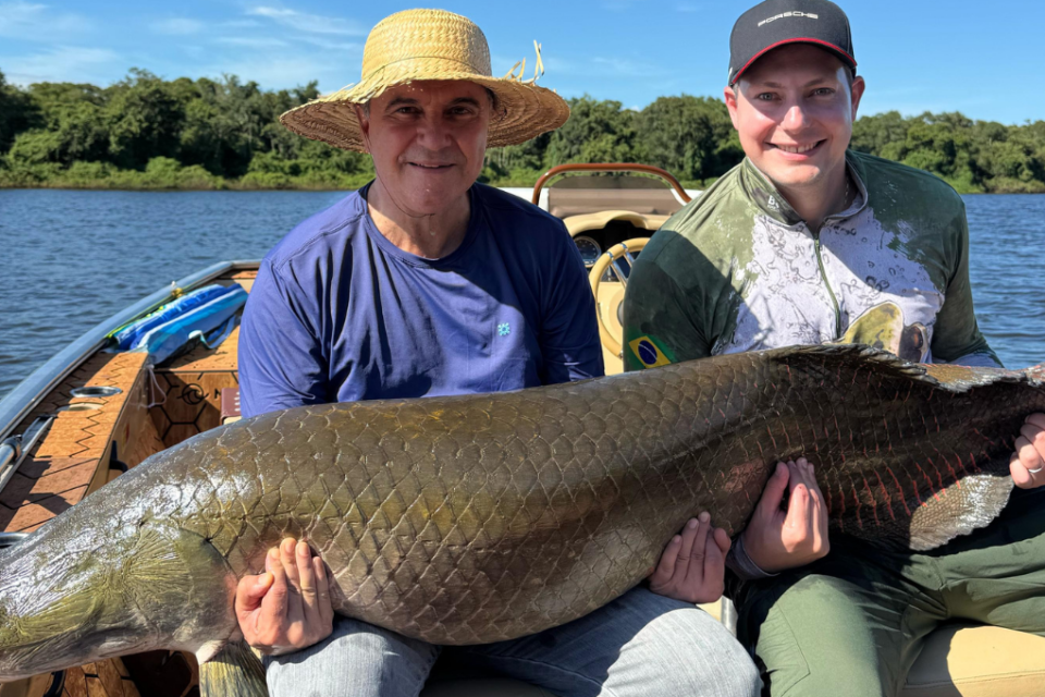 Imagem colorida mostra dois homens segurando um peixe gigante, pescado no Rio Araguaia.
