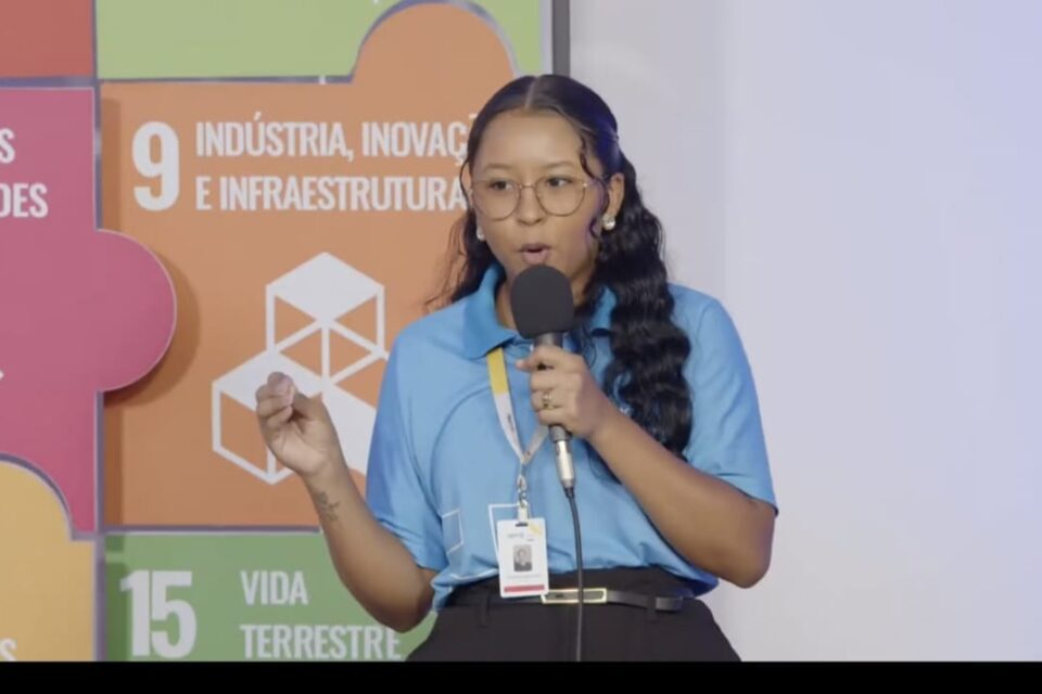 Mulher jovem de cabelos longos ministra palestra e segura um microfone na mão