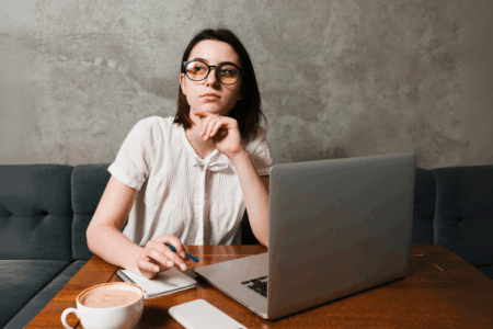 Mulher jovem, de óculos, setada em uma mesa, em frente ao laptop.