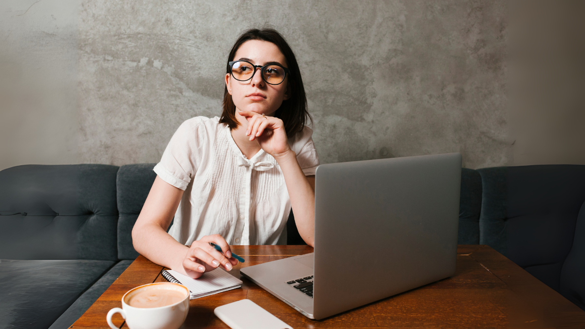 Mulher jovem, de óculos, setada em uma mesa, em frente ao laptop.