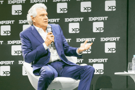 Homem grisalho vestindo um terno, sentado em uma cadeira branca segurando um microfone e gesticulando