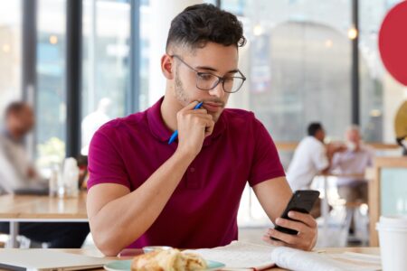 Homem jovem, de óculos, olha para o celular e segura uma caneta. Ele está sentado em uma mesa com papéis.
