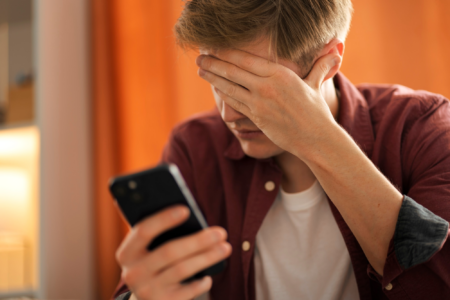 Homem jovem triste, com a mão no rosto e olhando para o celular