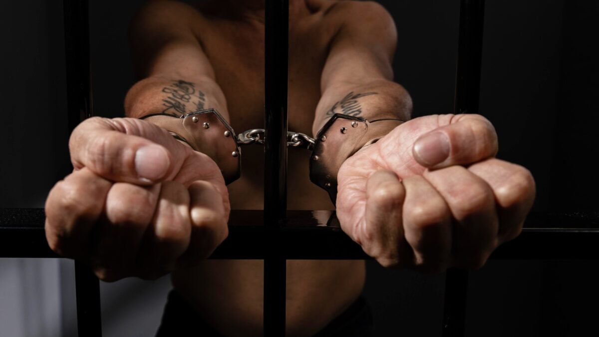 Foto ilustrativa mostra homem algemado e atrás das grades (Foto: reprodução/Freepik)