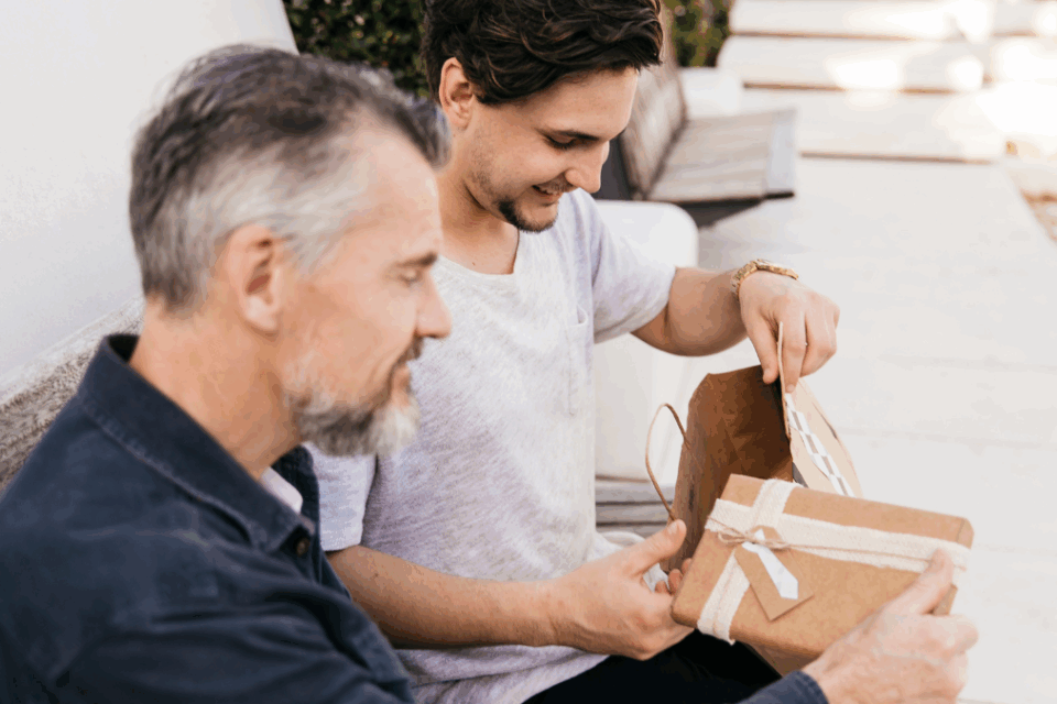 Dois homens sentados em um sofá lado a lado sorriem enquanto o mais jovem entrega um presente ao mais velho