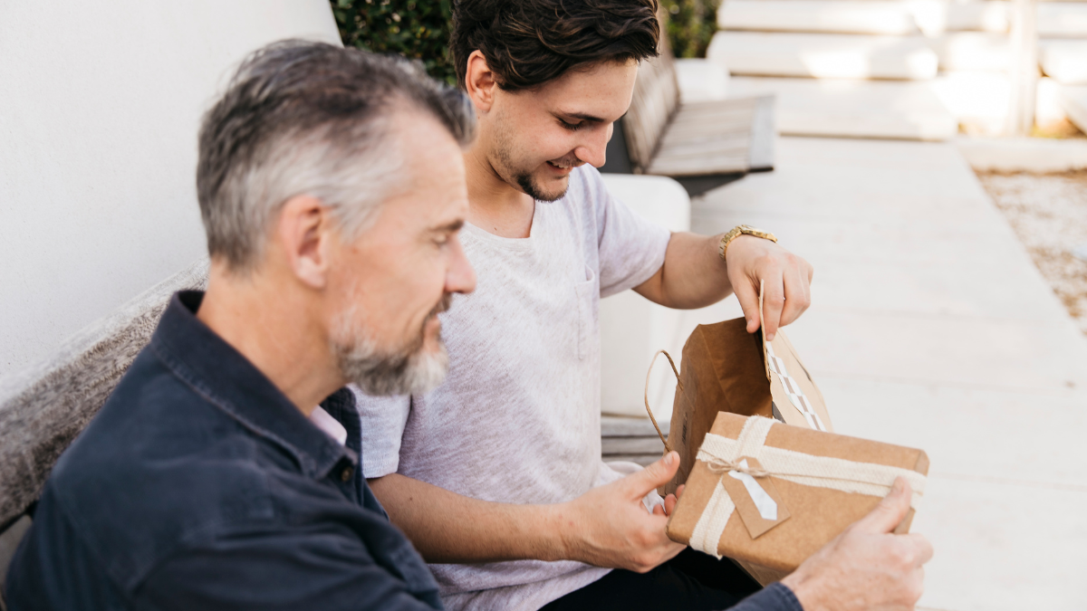 Dois homens sentados em um sofá lado a lado sorriem enquanto o mais jovem entrega um presente ao mais velho