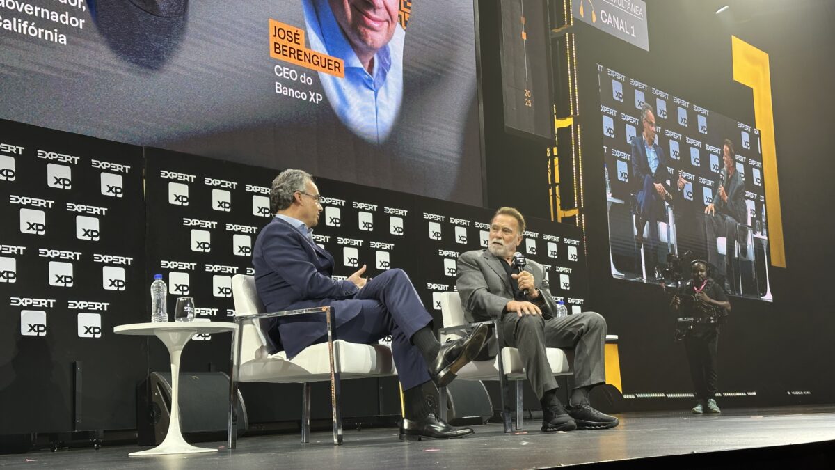 Dois homens estão sentados em um palco com microfones nas mãos. Ao fundo, um painel preto com telão de led com a foto deles.