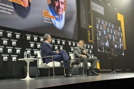 Dois homens estão sentados em um palco com microfones nas mãos. Ao fundo, um painel preto com telão de led com a foto deles.