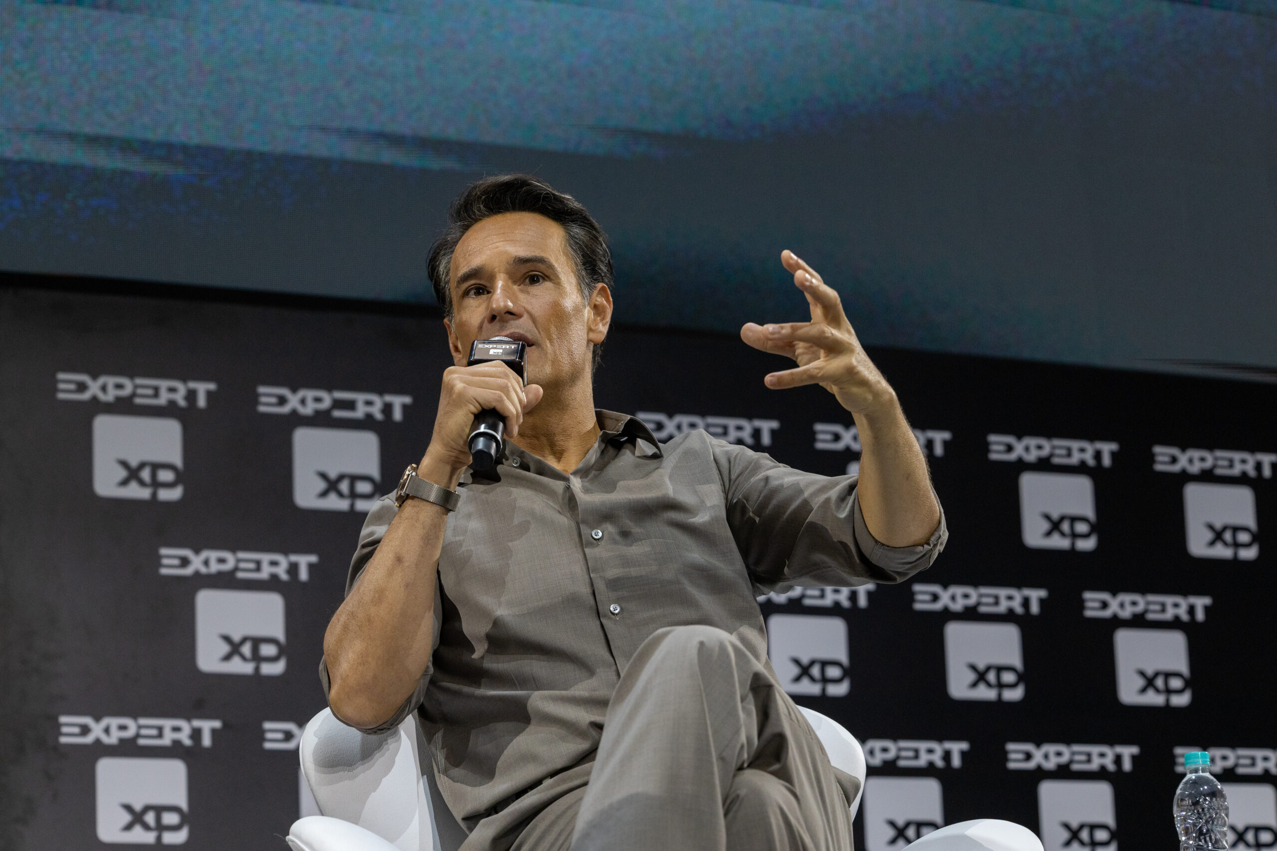 Homem gesticula e segura um microfone, ele está sentado. Ao fundo, o painel escuro com a logomarca do evento
