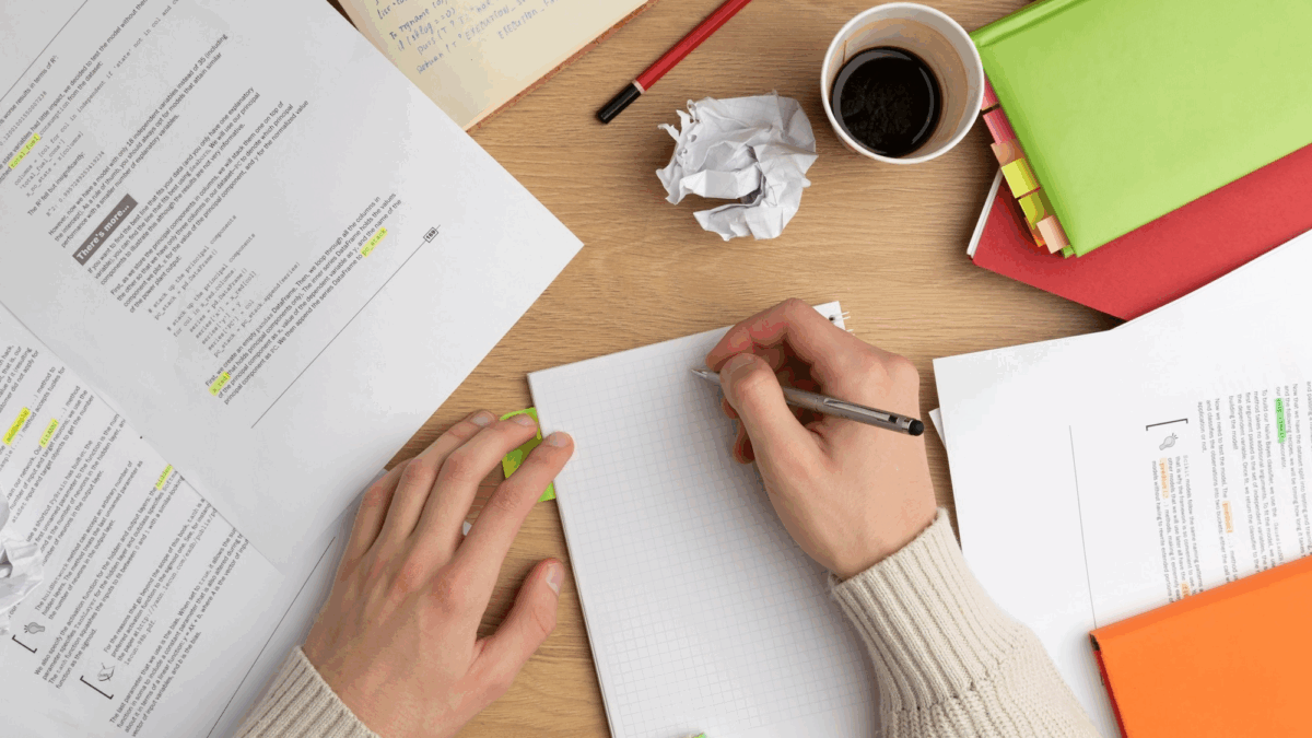 Mesa com papeis, copo de café e anotações. Em primeiro plano, duas mãos, uma delas escreve no papel