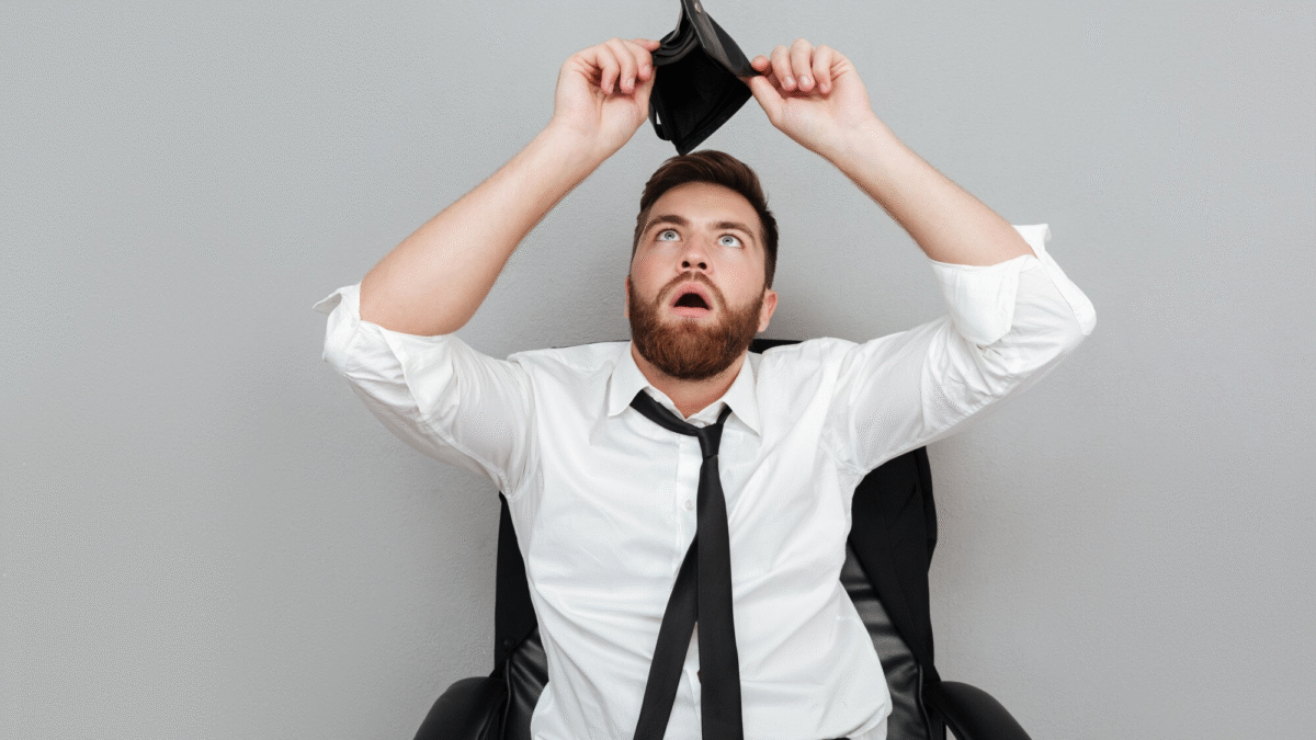 Homem de barba e bigode olha para sua carteira vazia acima de sua cabeça. Ele veste camisa branca, gravata e calça pretas e está sentado em uma cadeira de escritório