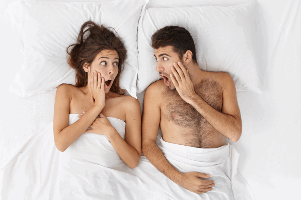 Mulher e homem deitados em uma cama com lençóis brancos e com expressão surpresa