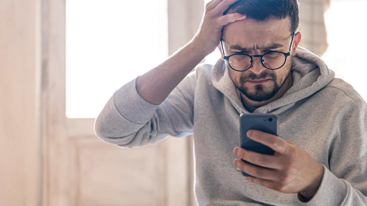 Homem com óculos e a mão no rosto. Ele segura um celular e tem expressão preocupada.