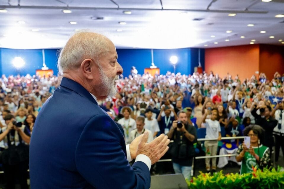 Imagem mostra presidente Lula durante solenidade do Governo Federal. Ao fundo, plateia acompanha evento em auditório cheio