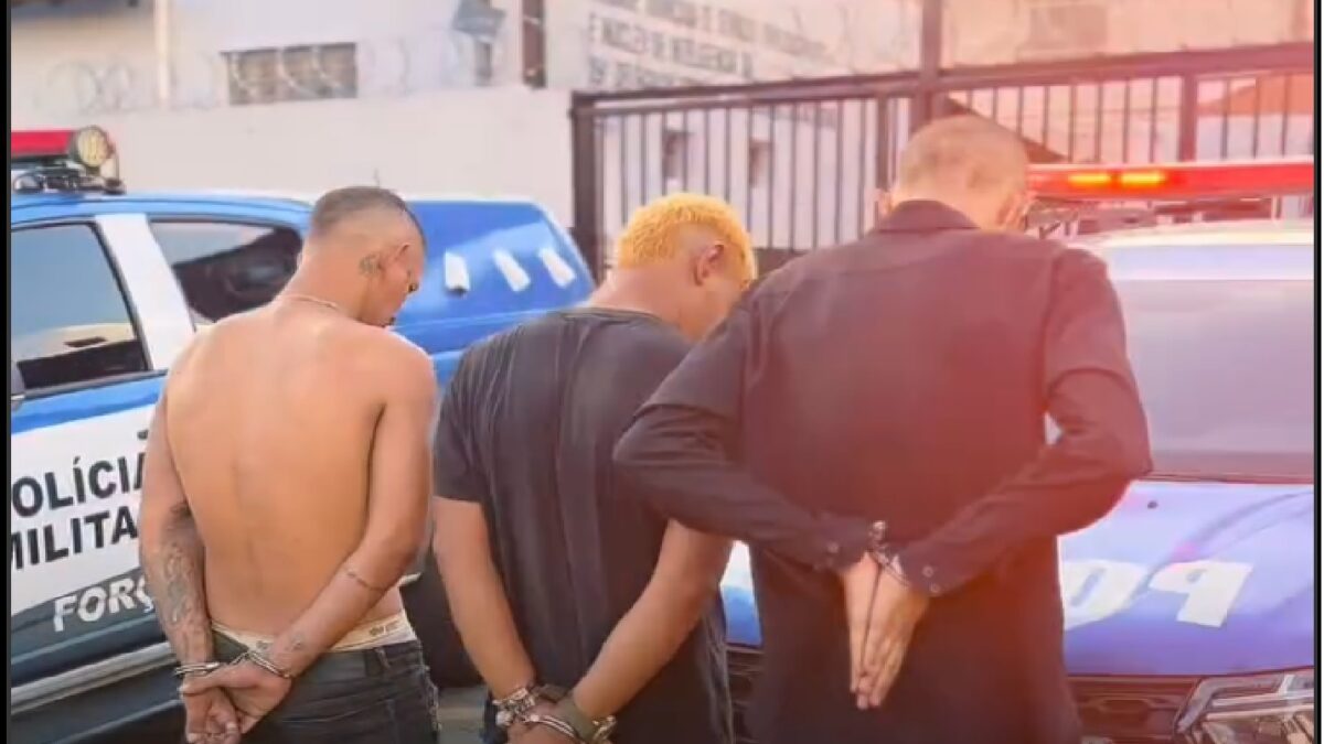 Imagem mostra três homens detidos e posicionados em frente a uma viatura da Polícia Militar de Goiás