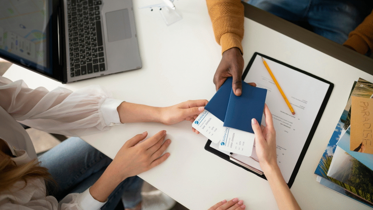Mãos de três pessoas seguram dois passaportes com passagens dentro sobre uma mesa. A mesa é branca e tem itens de escritório e um notebook