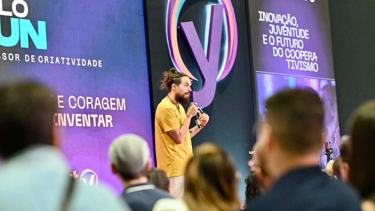 Murilo Gun palestrando na Feira do Empreendedor. Ele é um homem de camisa amarela, de cabelos longos e amarrados e que segura um microfone. Ao fundo, o painel do evento e, em primeiro plano, pessoas assistem à palestra.