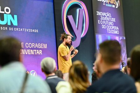 Murilo Gun palestrando na Feira do Empreendedor. Ele é um homem de camisa amarela, de cabelos longos e amarrados e que segura um microfone. Ao fundo, o painel do evento e, em primeiro plano, pessoas assistem à palestra.