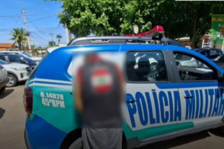 Imagem mostra suspeito algemado ao lado de viatura da polícia