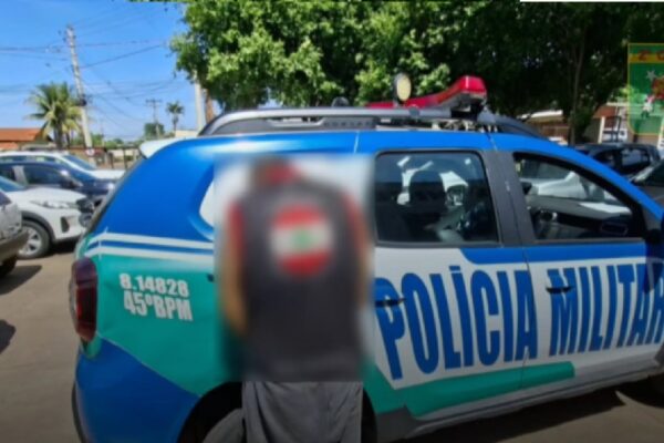 Imagem mostra suspeito algemado ao lado de viatura da polícia