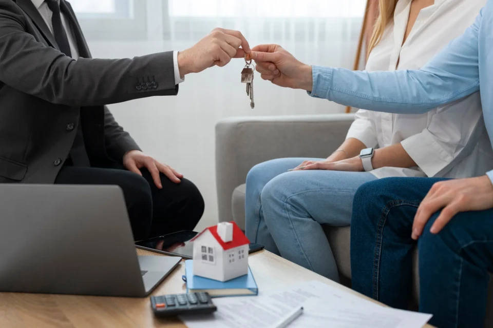 Homem de terno entreva uma chave a outro homem, que está ao lado de uma mulher. Os três estão sentados diante de uma mesa de centro com uma escultura de casa.