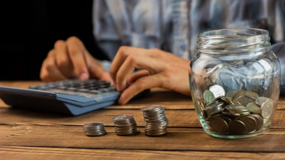 Mãos de um homem usando uma calculadora. Na mesa, um pote de vidro com moedas e três pilhas de moedas.