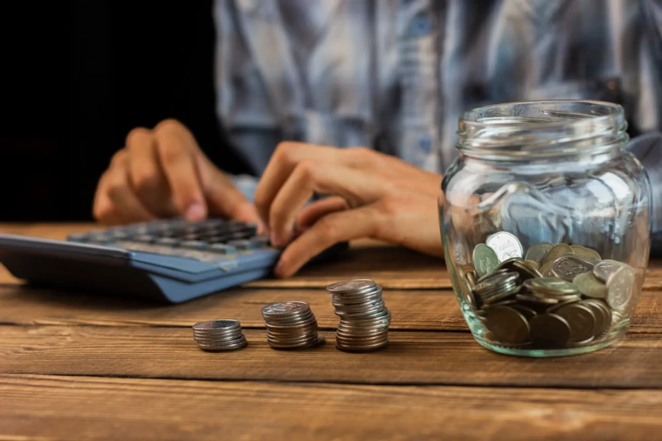 Mãos de um homem usando uma calculadora. Na mesa, um pote de vidro com moedas e três pilhas de moedas.