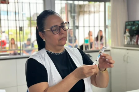 Enfermeira prepara uma injeção. Ela é uma mulher com cabelos escuros, presos, usa óculos, veste uma camiseta preta e um colete branco