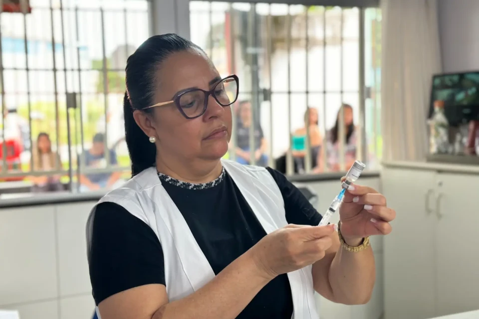 Enfermeira prepara uma injeção. Ela é uma mulher com cabelos escuros, presos, usa óculos, veste uma camiseta preta e um colete branco