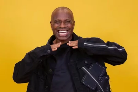 Homem negro sorri. Ele veste um casaco escuro e é careca