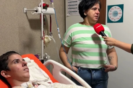 Duas mulheres. Uma deitada em uma cama hospitalar e outra, em pé, sendo entrevistada