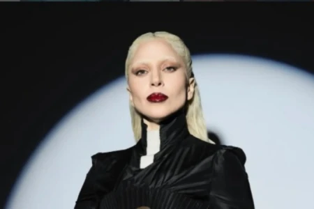Lady Gaga em close. Ela é uma mulher de cabelos claros, veste um vestido preto e maquiagem carregada.