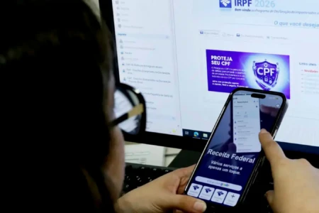 Mulher em frente a uma tela de computador com o celular na mão