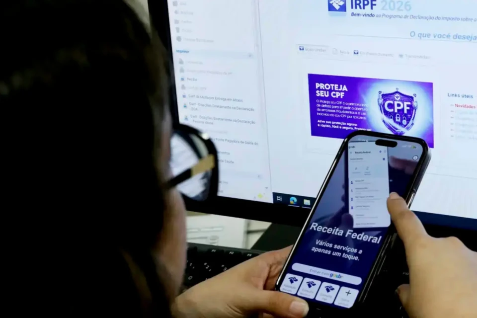 Mulher em frente a uma tela de computador com o celular na mão