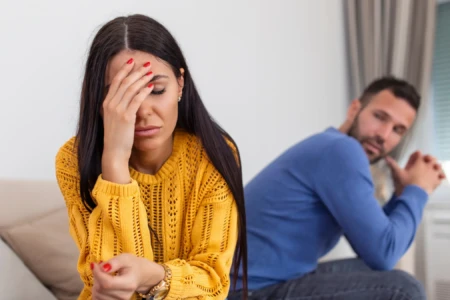Mulher triste de cabelos longos e escuros no primeiro plano e um homem em segundo plano olhando para ela