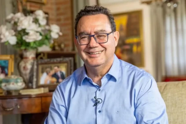 Homem branco com cabelos escuros e óculos. Veste uma camisa social azul clara