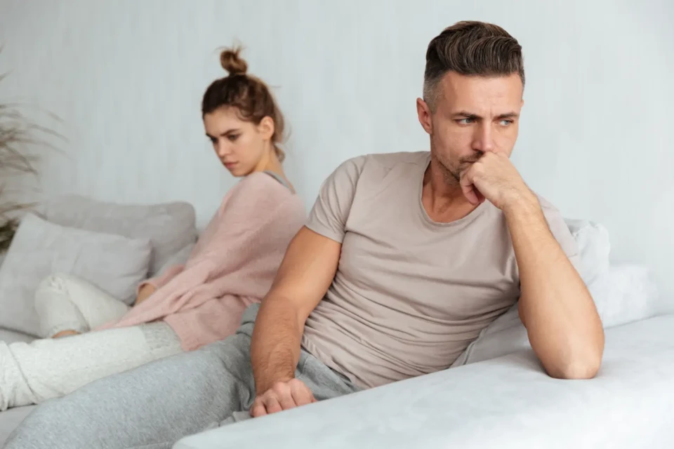 Homem em primeiro plano com expressão chateada. Mulher ao fundo, em posição desconfortável