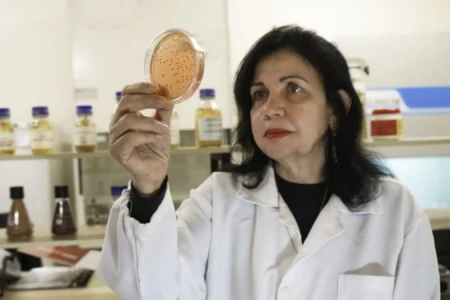 Mulher com cabelos pretos segura amostra. Ela está de jaleco em um laboratório