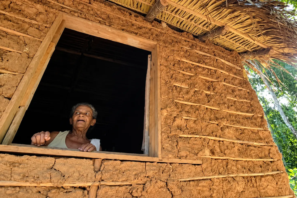 Moradora de comunidade quilombola observa o lado de fora pela janela de casa construída com barro e madeira (Foto: Reprodução/YouTube)
